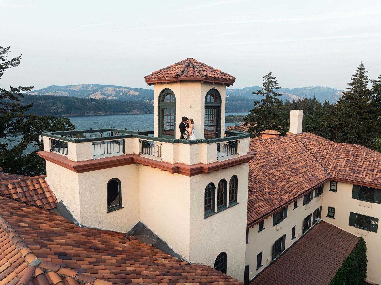 Couple embracing atop a Mediterranean-style villa tower with scenic lake and mountain views at sunset.