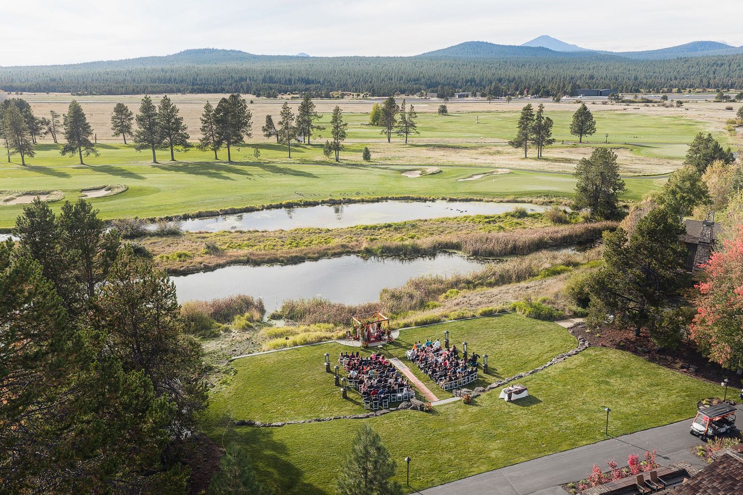 Aerial view of an outdoor wedding ceremony on a golf course with a pond, pine trees, and mountain backdrop.