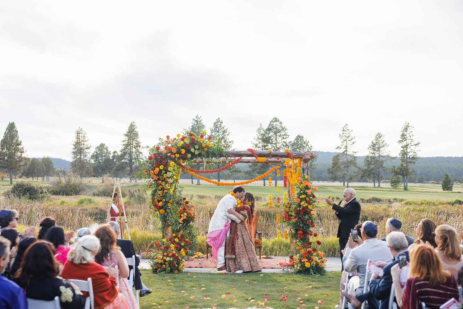 Outdoor Indian wedding ceremony under colorful floral arch with couple exchanging vows on green lawn.