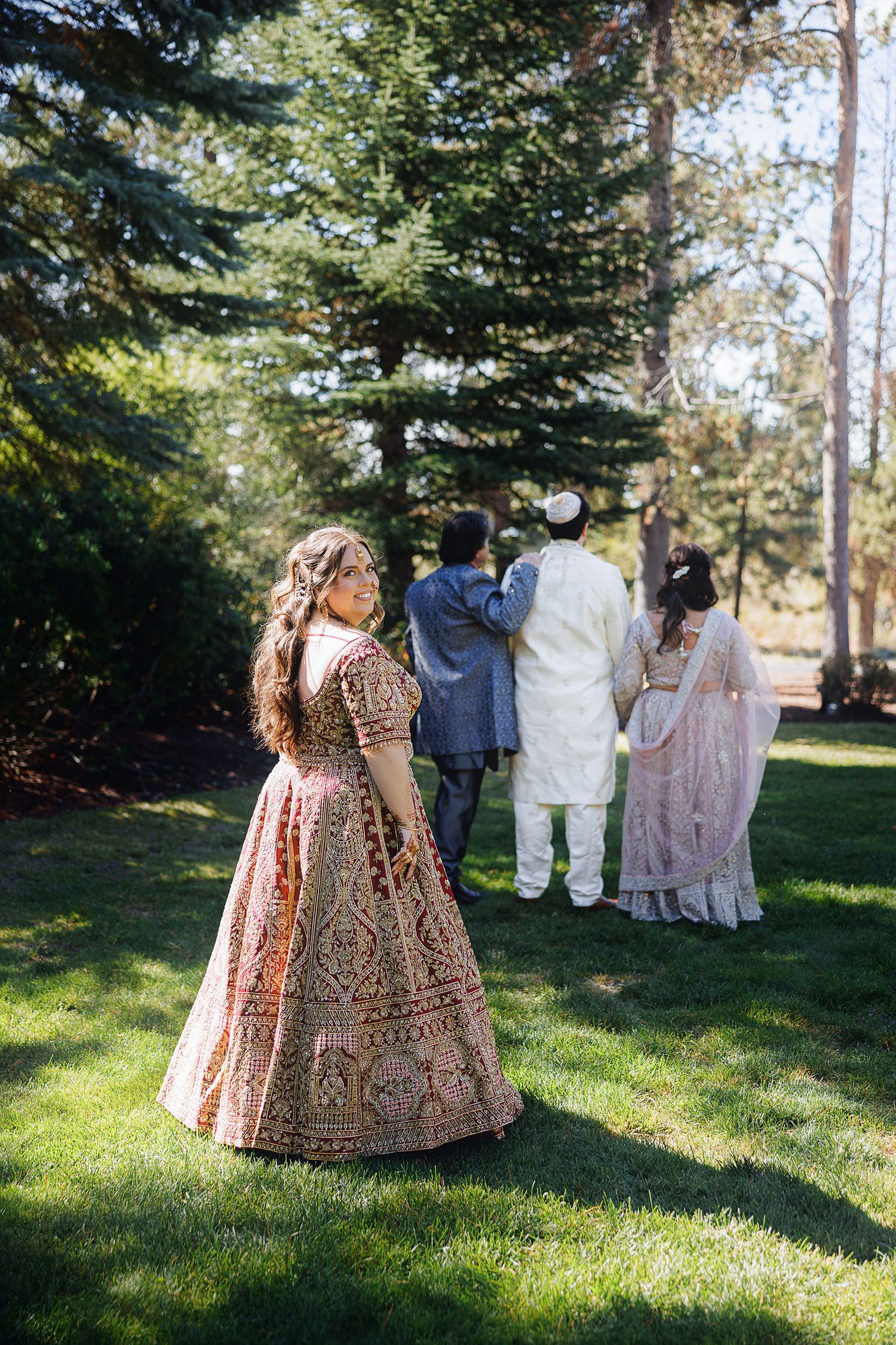 Woman in ornate pink lehenga stands on lawn as wedding guests in traditional Indian attire gather among tall trees.