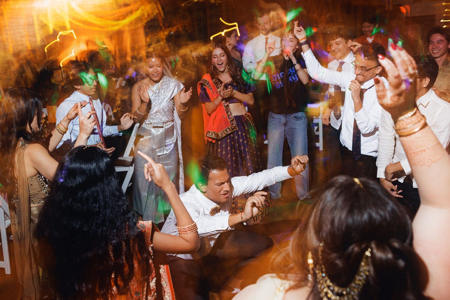 Guests dancing energetically at a lively indoor party or wedding reception with colorful lights.
