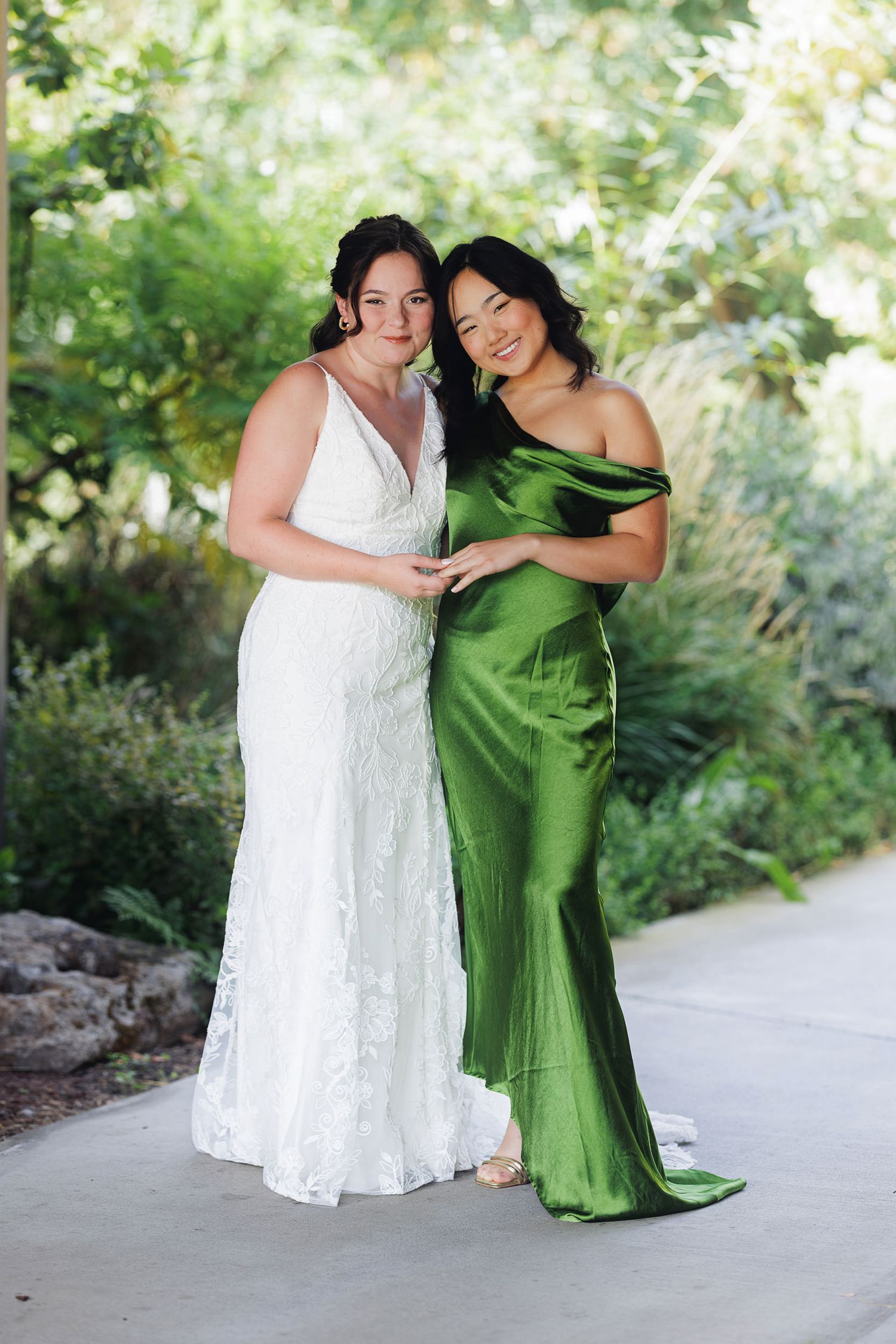 Bride in white lace gown poses with bridesmaid in emerald green satin dress outdoors.