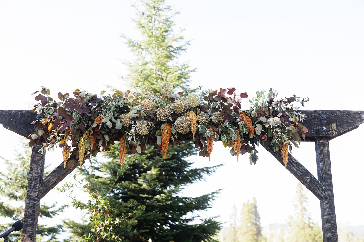 Rustic wooden wedding arch adorned with lush autumn floral garland featuring dried oranges, greenery, and blooms against evergreen trees.