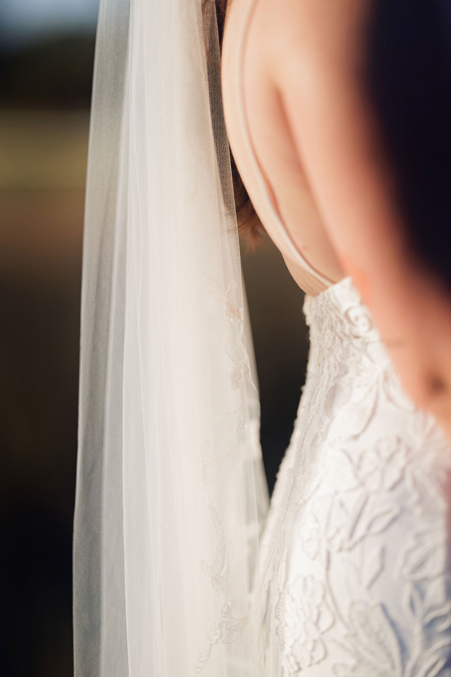 Close-up of a bride in a backless lace wedding gown with a flowing white veil draped elegantly over her shoulder.