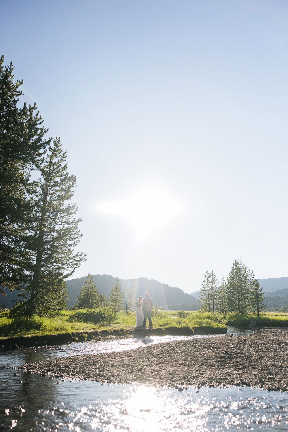 Silhouettes of a couple stand against a bright mountain landscape with sunlight sparkling on water.