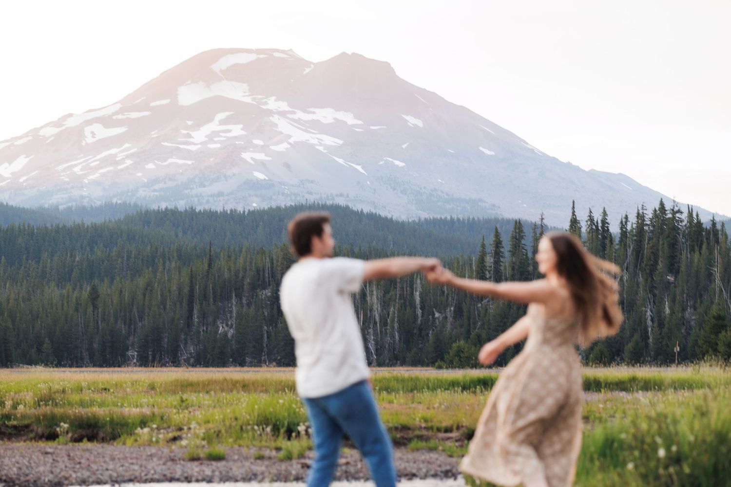 A couple dances in a scenic mountain meadow surrounded by evergreen forest and snowy peaks in the distance.