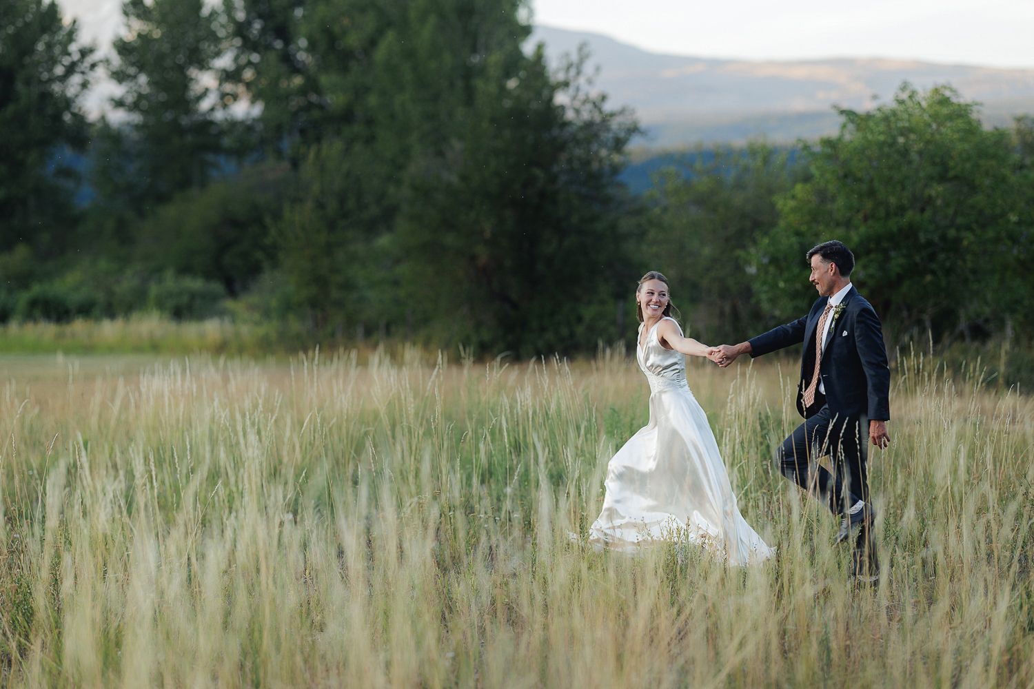 A couple in wedding attire stands together in a scenic field with mountains and trees in the background.
