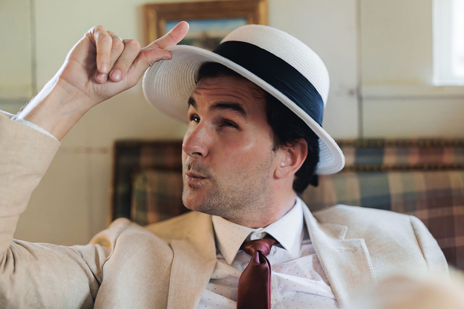 Someone wearing a white fedora hat, white dress shirt and red tie relaxes on a couch while adjusting their hat.