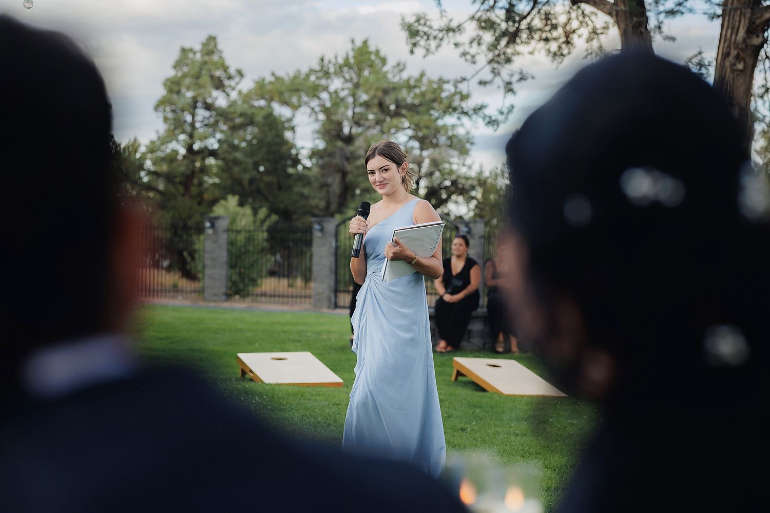 A bridesmaid in a light blue dress walks through a garden setting while guests in dark attire watch.