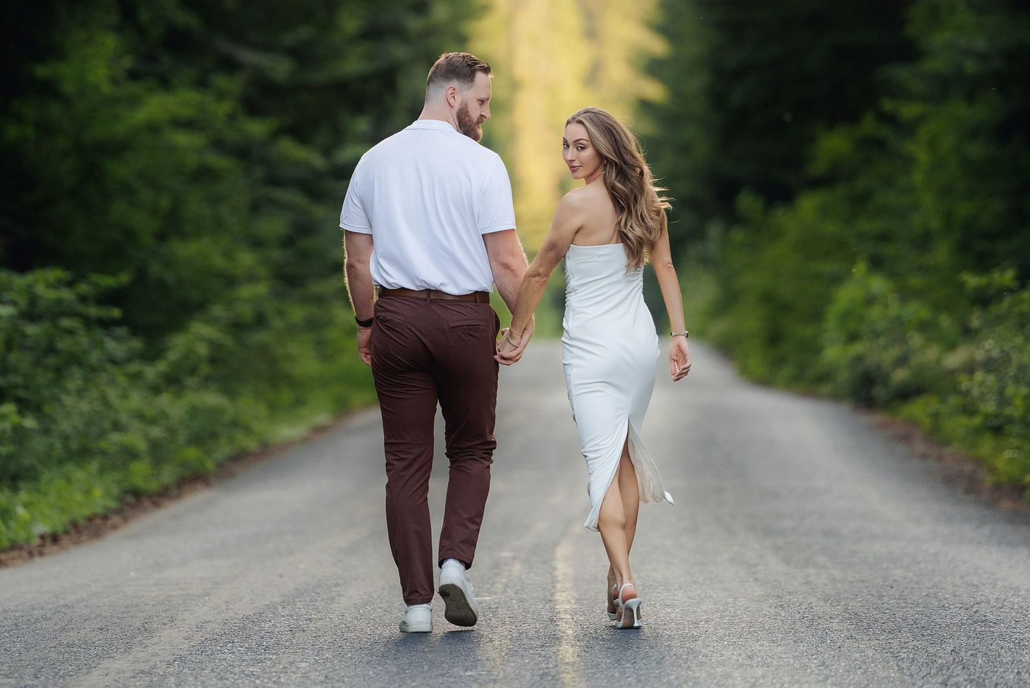 A romantic couple walks together on a tree-lined dirt road, the bride in a white dress and groom in brown pants.