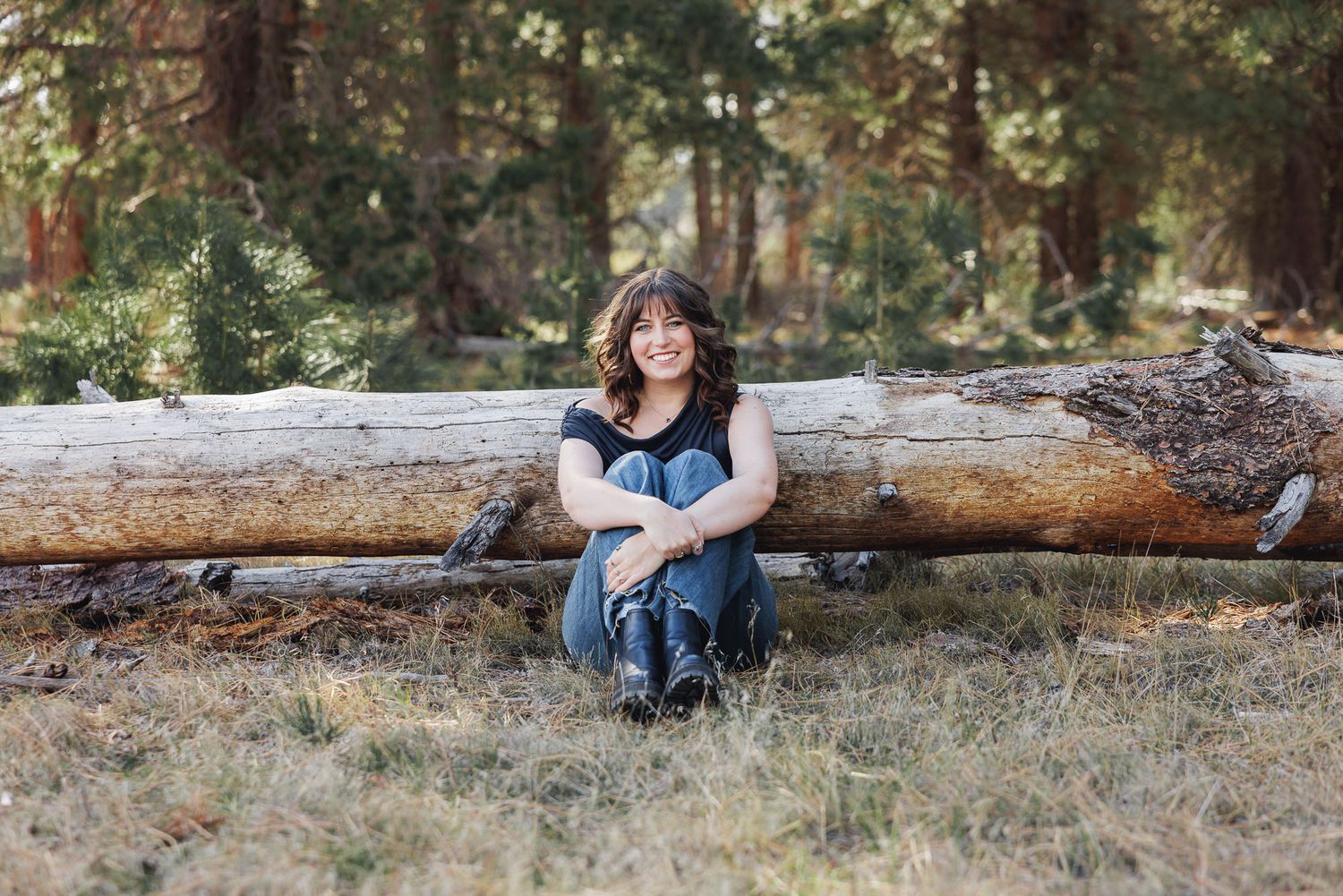 Young woman with curly hair sits smiling against a fallen log in a sunlit pine forest wearing jeans and a black top.