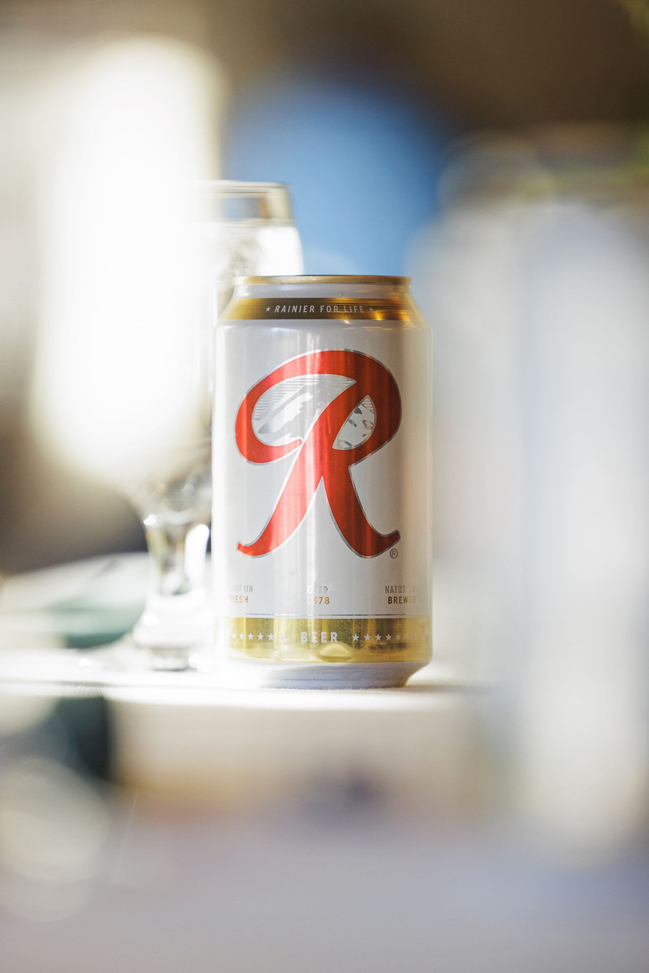 A can of Rainier Beer with bold red R logo sits on a table surrounded by blurred glassware in soft light.