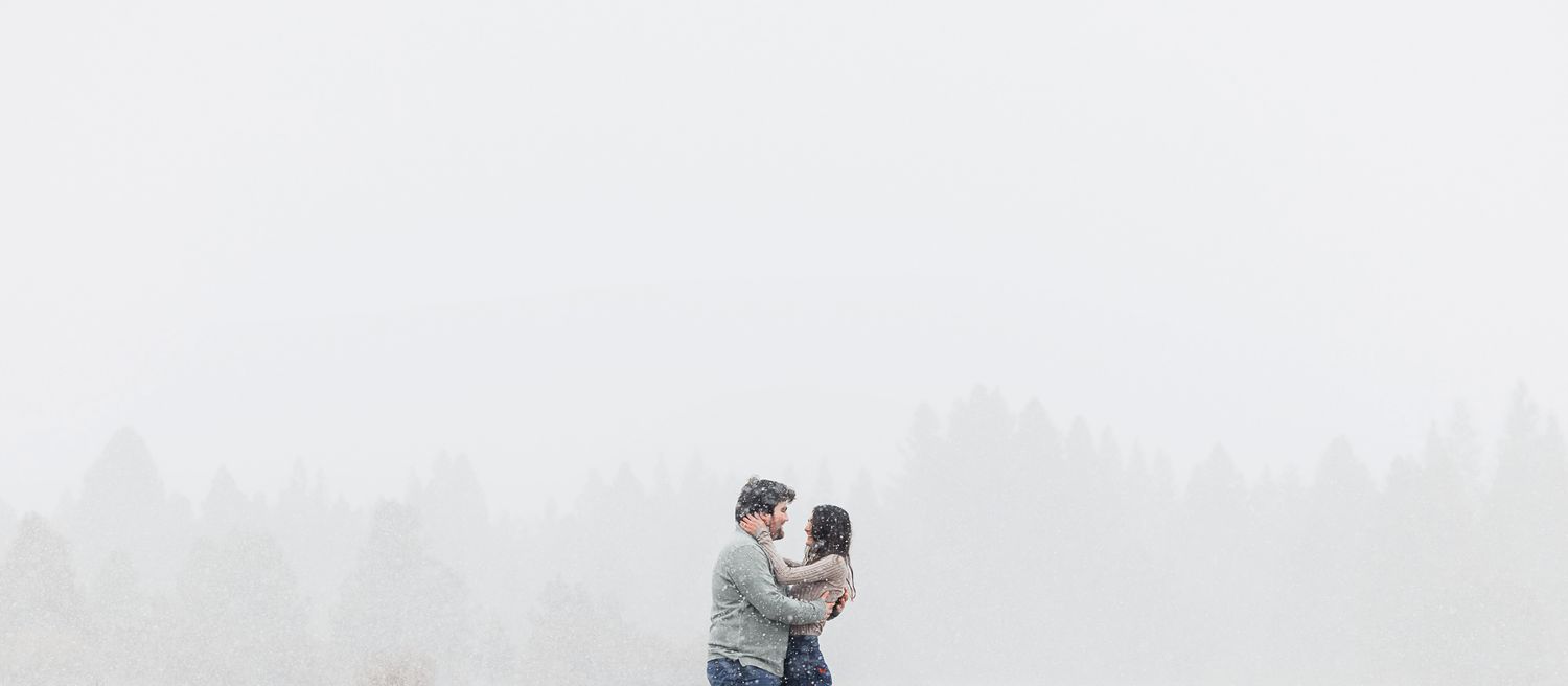 A couple embraces in a romantic moment against a misty white winter landscape.