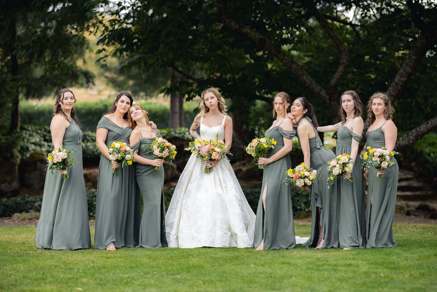 Wedding party in sage green bridesmaid dresses and white bridal gown pose with yellow sunflower bouquets on lawn.