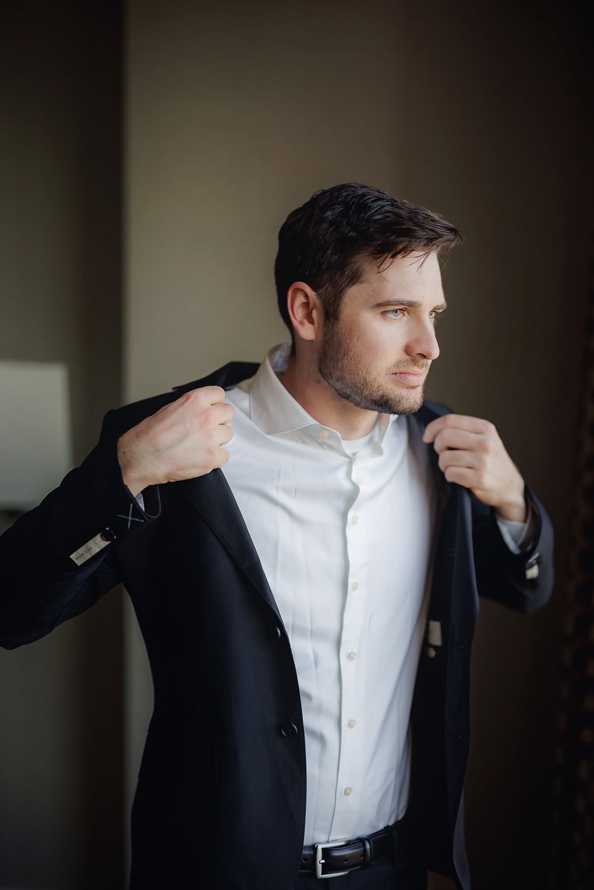 Man in white dress shirt putting on a black suit jacket while looking to the side in natural lighting.