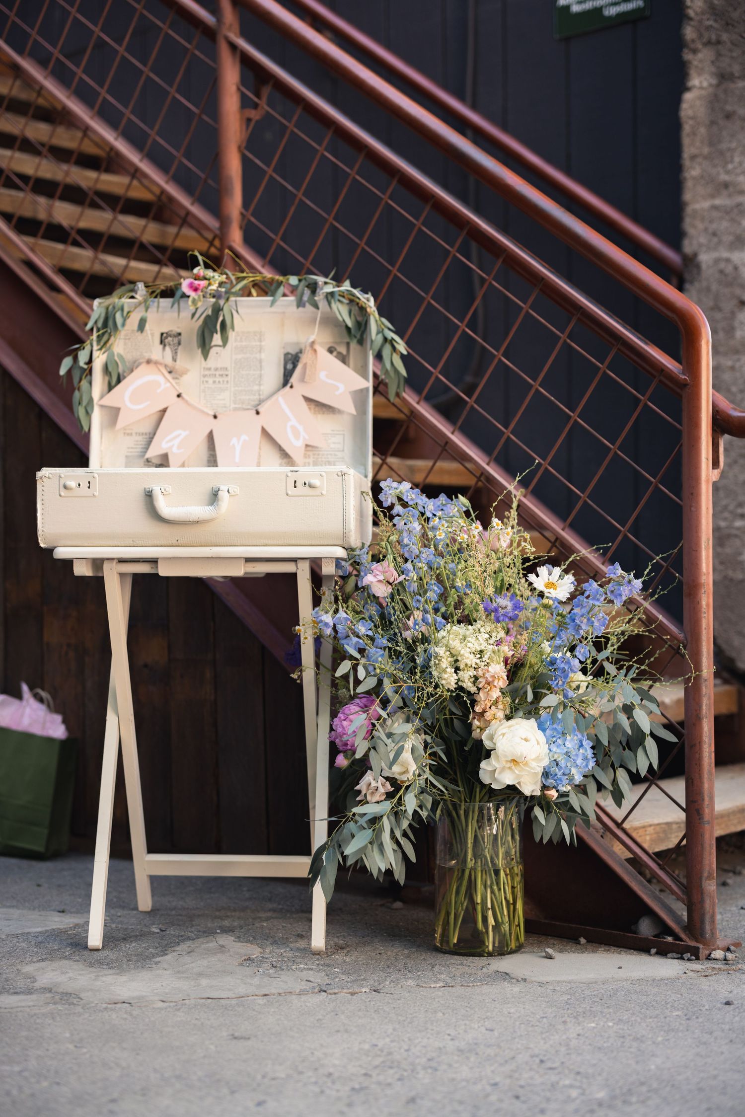 Vintage white suitcase on stand with floral decorations next to large rustic bouquet of blue and white flowers by metal stairs.