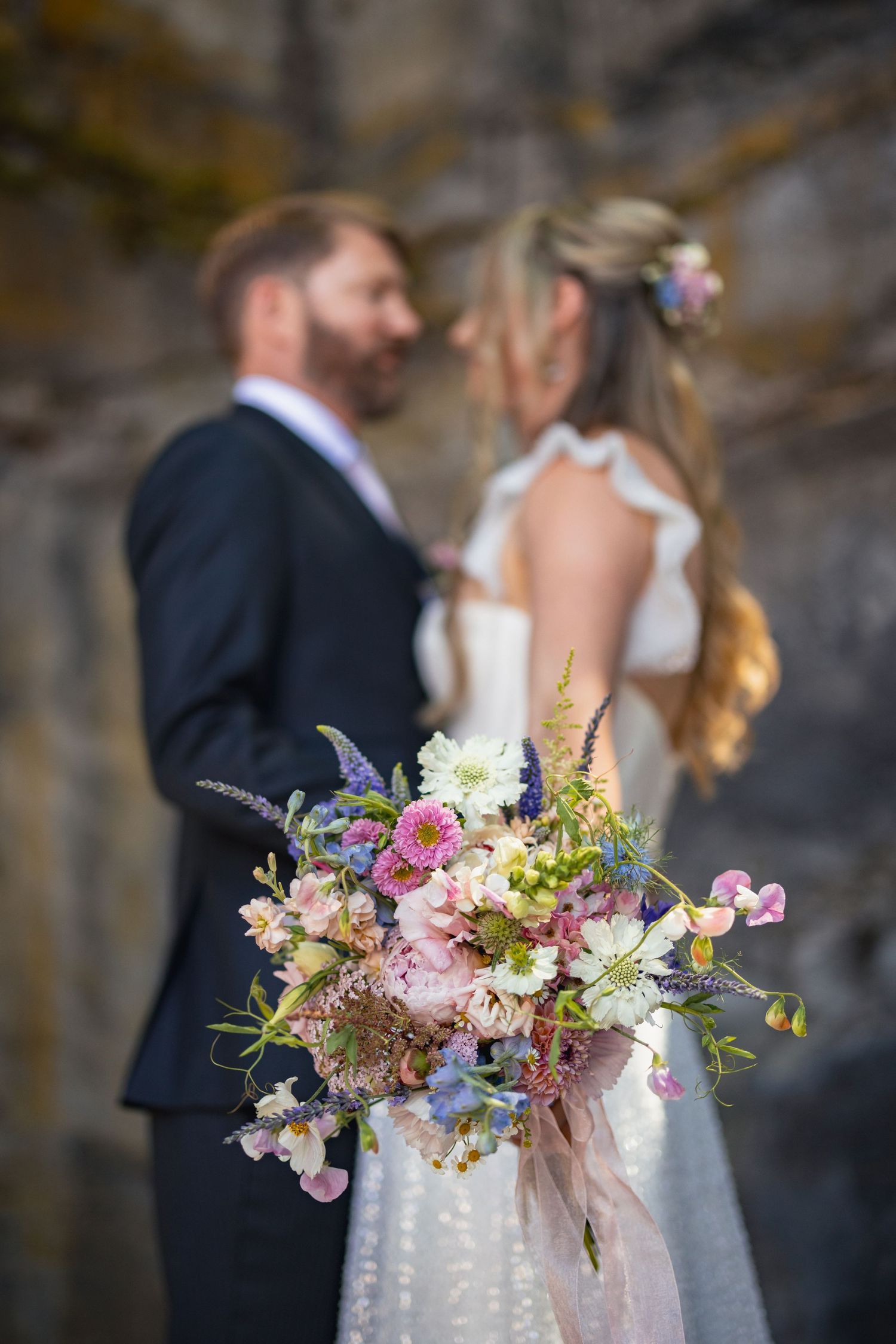 A romantic wedding moment featuring a beautiful spring bouquet with pink, purple and yellow wildflowers in the foreground.