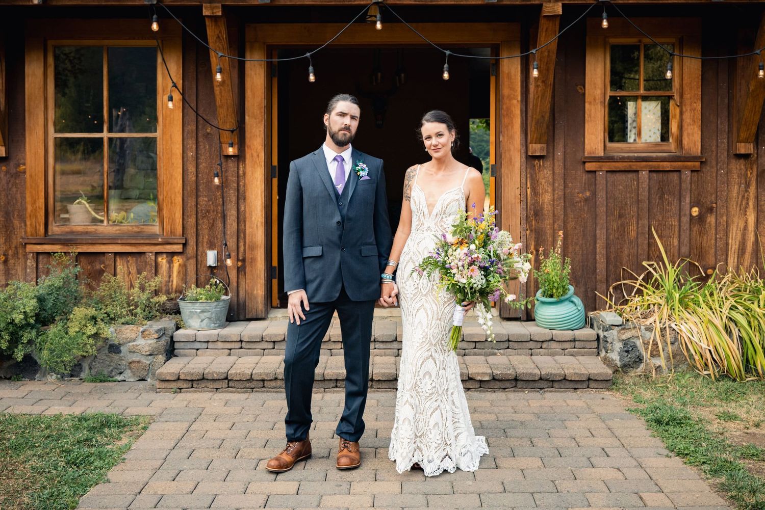 A couple in wedding attire stands in front of a rustic wooden building entrance with stone steps and garden plants.