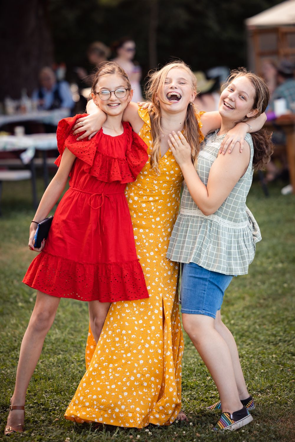 Three friends in colorful dresses share a candid laughing moment together outdoors on a summer day.