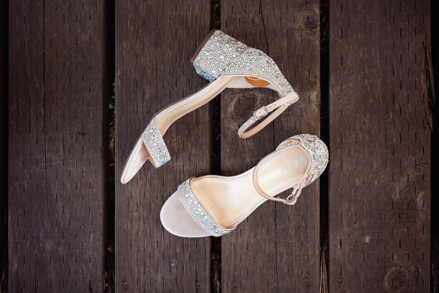 Elegant white bridal heels with sparkly rhinestone details displayed on dark wooden floorboards.
