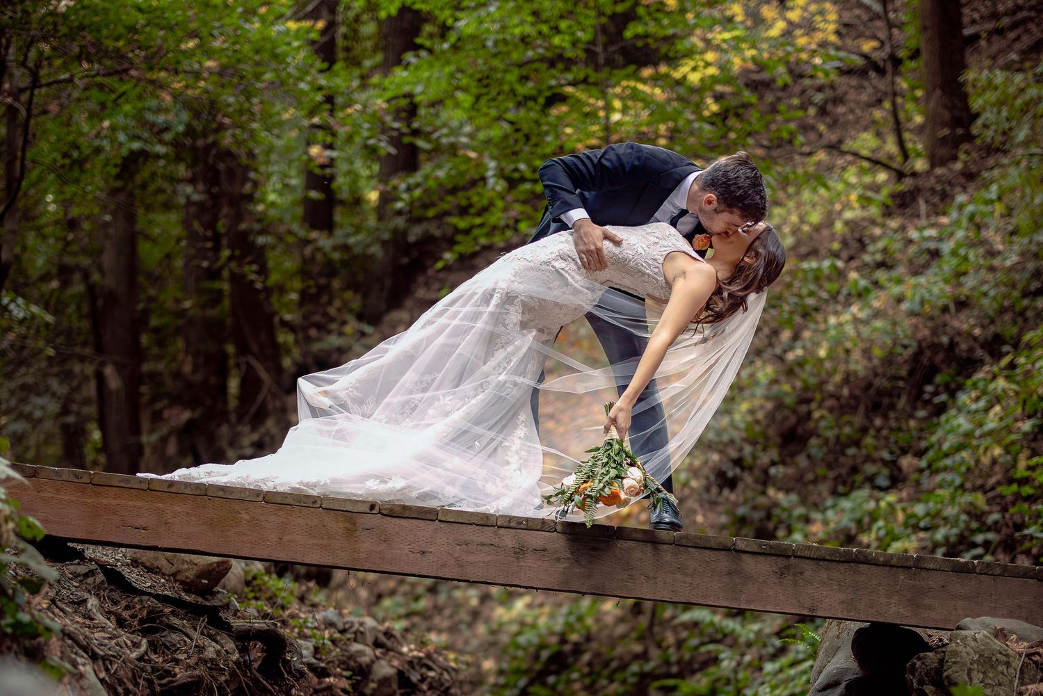 A romantic wedding couple snuggle on a rustic wooden bridge in a lush forest setting with flowing bridal veil.