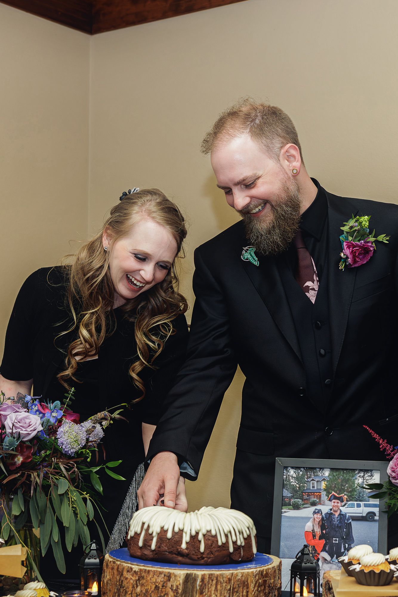 Smiling couple cutting a bundt wedding cake on a wood stump, groom in black suit with floral boutonniere.