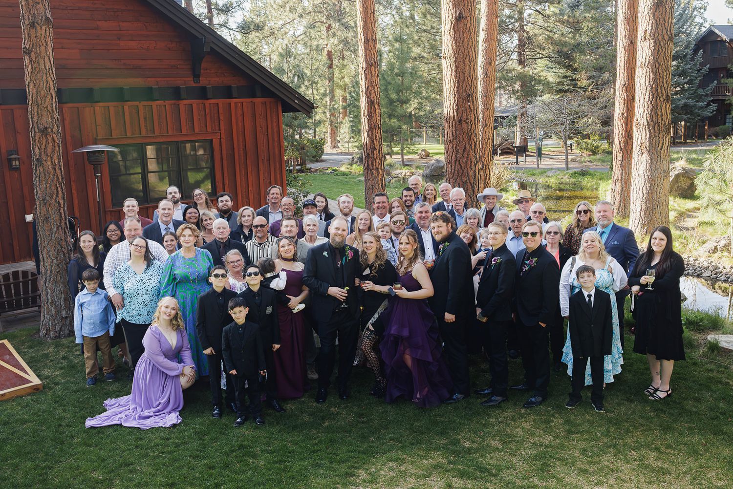 Large family group photo outdoors near rustic red cabin surrounded by pine trees, dressed in formal attire for a wedding.