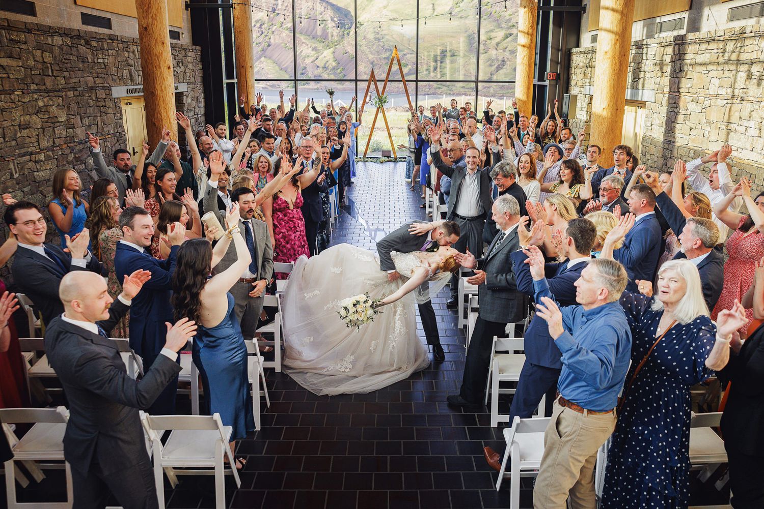Bride and groom share a kiss while wedding guests cheer in a stunning indoor ceremony venue.