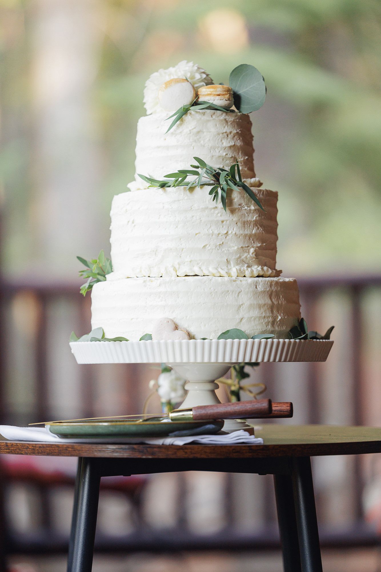 Elegant white wedding cake with textured frosting and delicate greenery accents displayed on a rustic wooden stand.