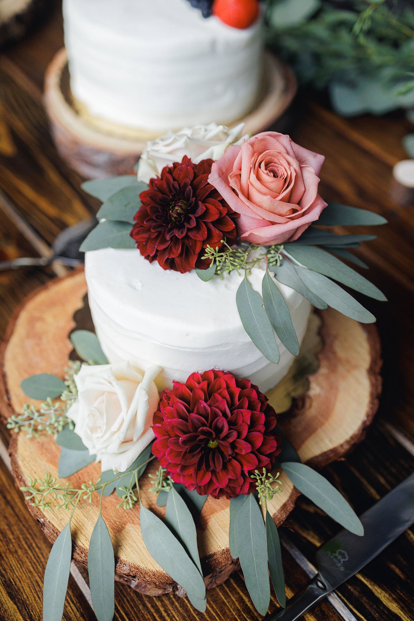 White wedding cake decorated with burgundy dahlias, pink roses and eucalyptus leaves on rustic wood slices.