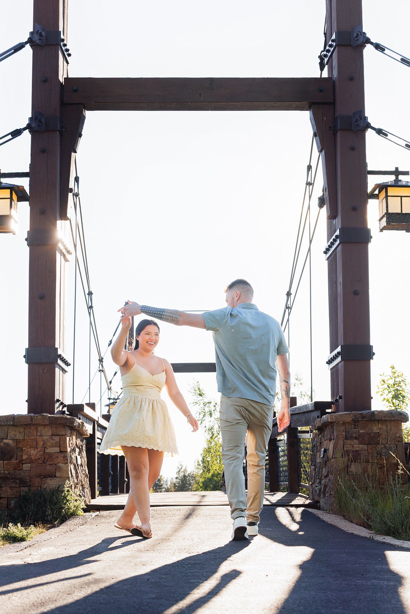 Couple dancing on a rustic suspension bridge at sunset, woman in yellow dress being twirled by man in blue shirt.