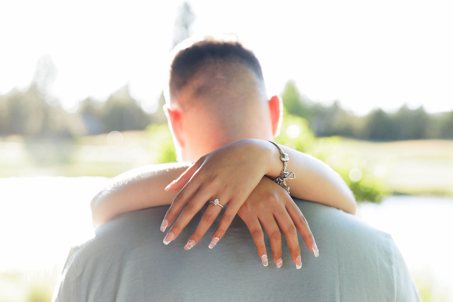 Woman with engagement ring embracing a man from behind outdoors in soft natural light.