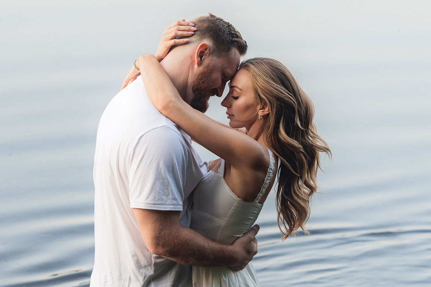 Two people embrace intimately near a calm waterfront as the breeze flows through their hair.