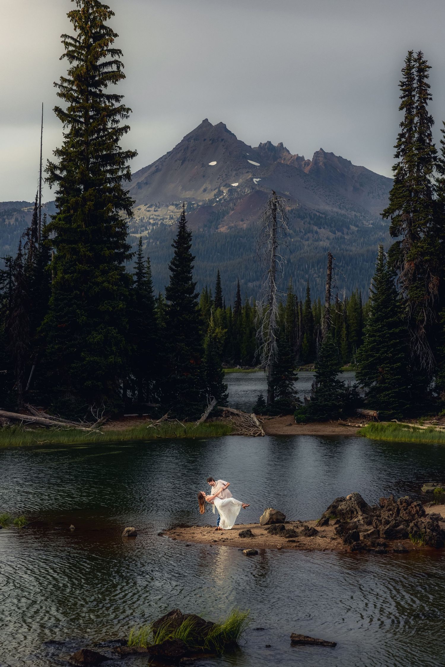 Mountain lake surrounded by evergreen trees with figures backlit by sunset creating silhouettes on the water's edge.