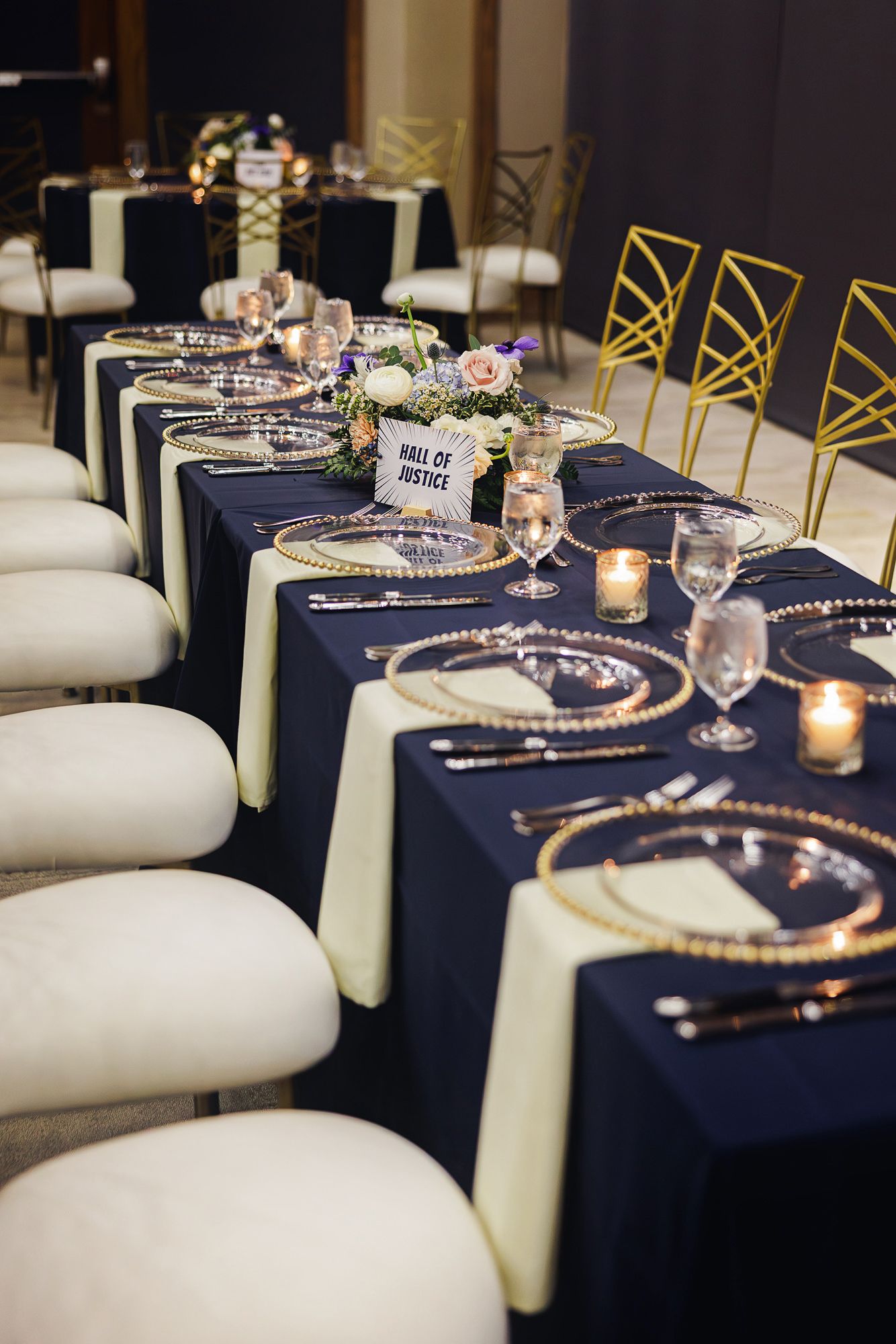 Long banquet table with navy linens, gold chargers, and white chairs set for an elegant wedding reception.