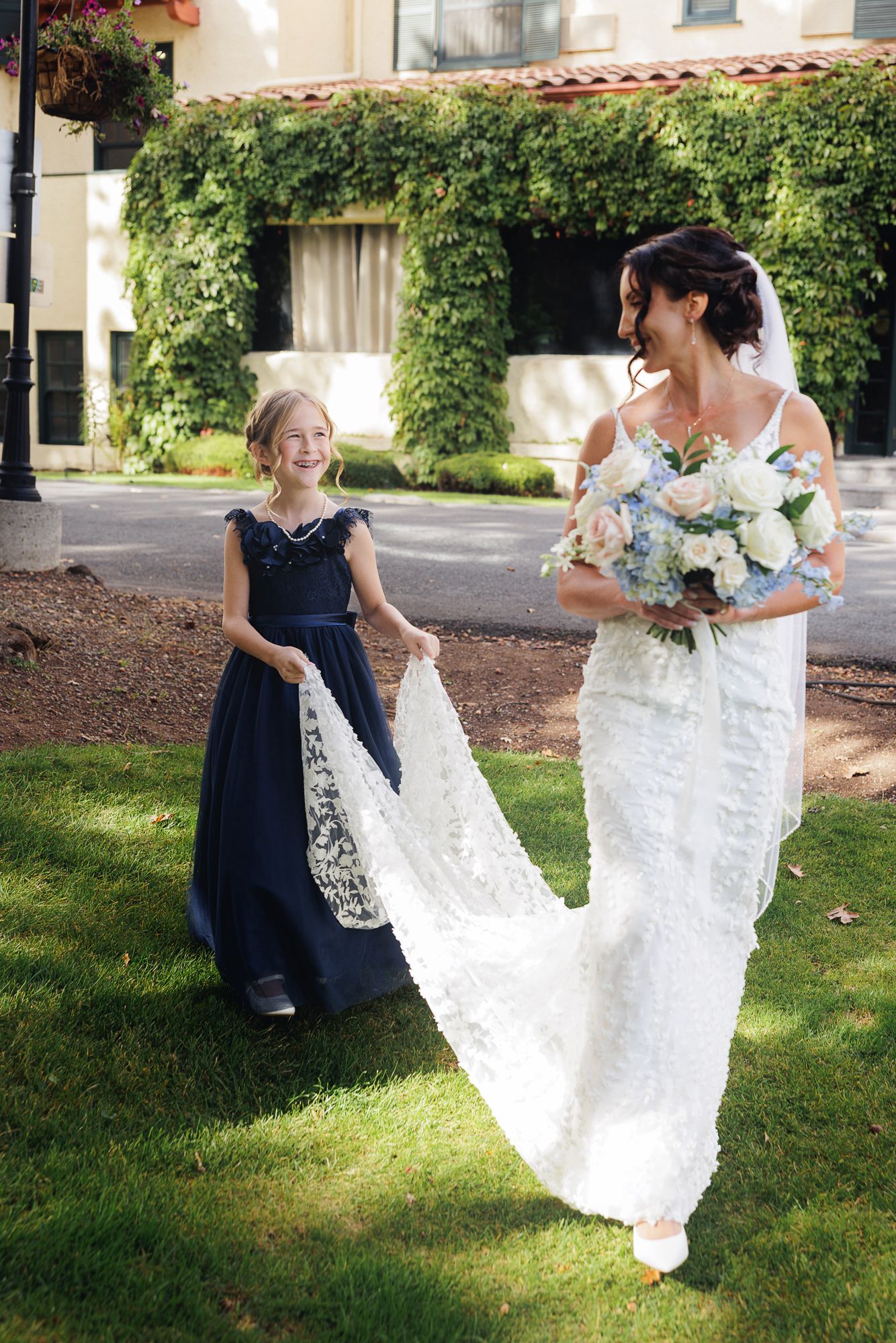 A bride in a white lace wedding dress twirls playfully on a grassy lawn with someone in a navy blue dress.