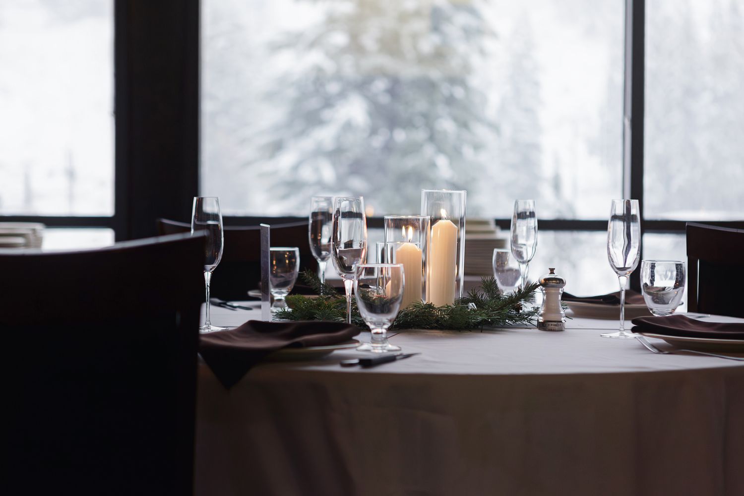 A rustic dining table set with white plates and wine glasses against a snowy winter backdrop through large windows.
