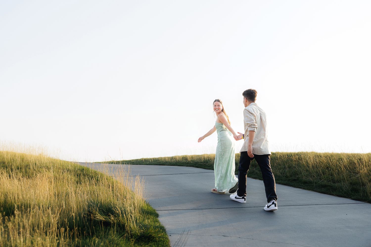 Couple holding hands walking on a path surrounded by green grass fields during golden hour.