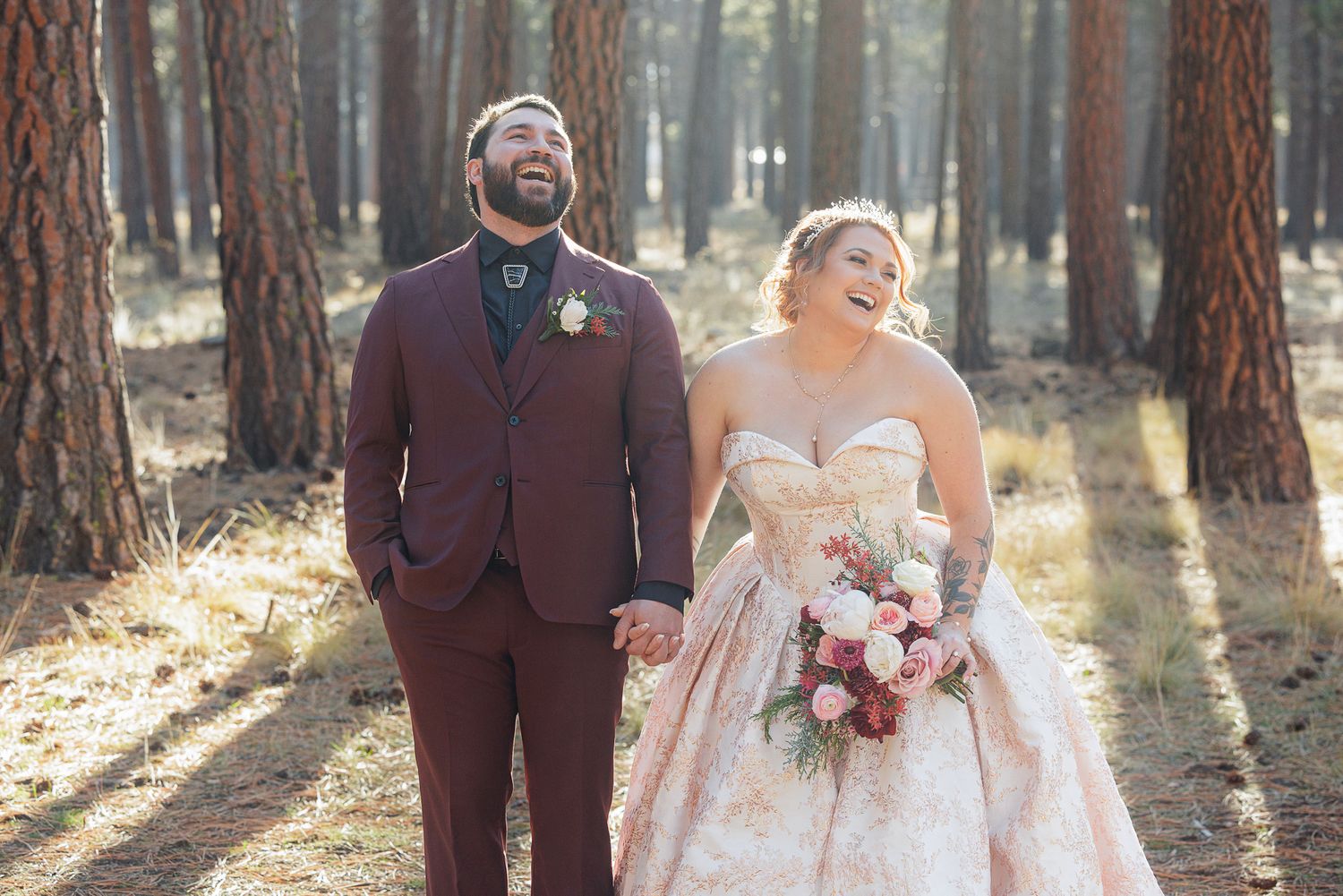 A joyful couple in wedding attire laughs together in a serene forest setting with sunlight filtering through trees.