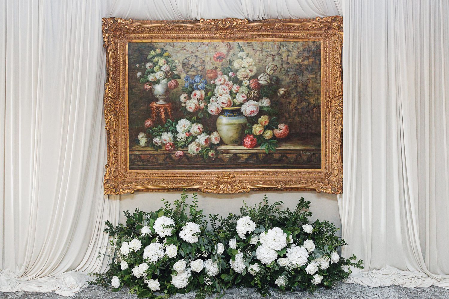 Gold-framed floral still life painting displayed above white flower arrangements against draped white fabric backdrop.