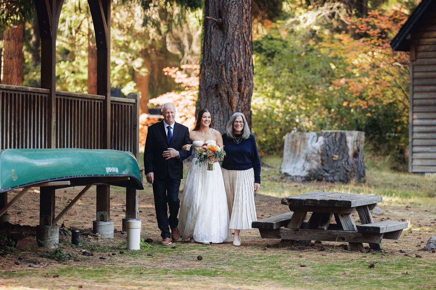 Bride with parents at outdoor wedding venue near rustic cabin, canoe, and picnic table surrounded by tall trees.
