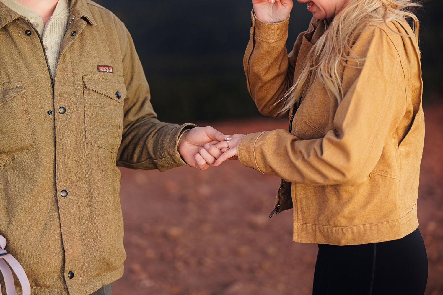 Couple in tan utility jackets holding hands outdoors against a red desert landscape, a romantic proposal or engagement moment.