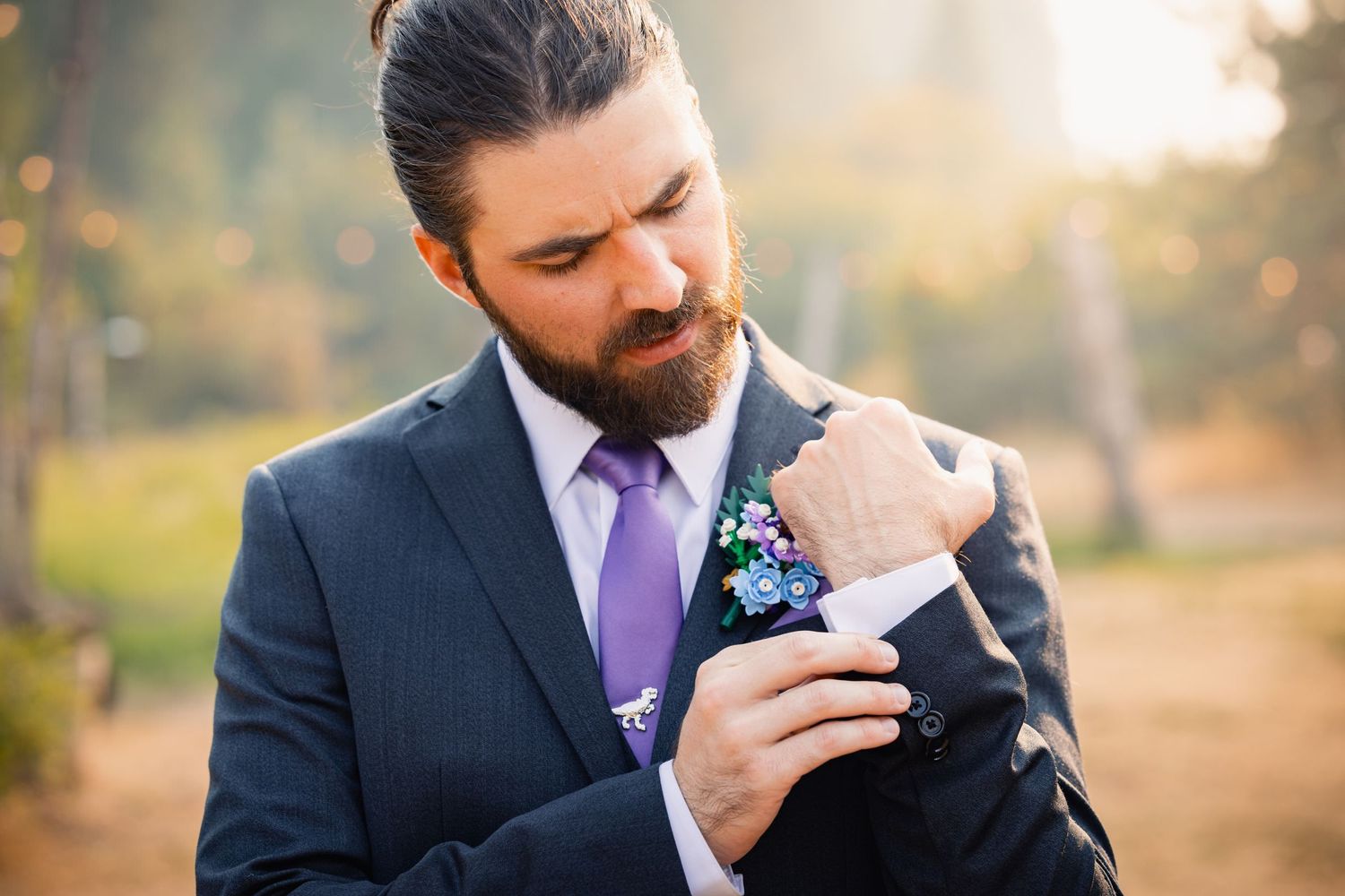 A groom in a navy suit with purple tie adjusts his boutonniere while standing outdoors in warm golden light.
