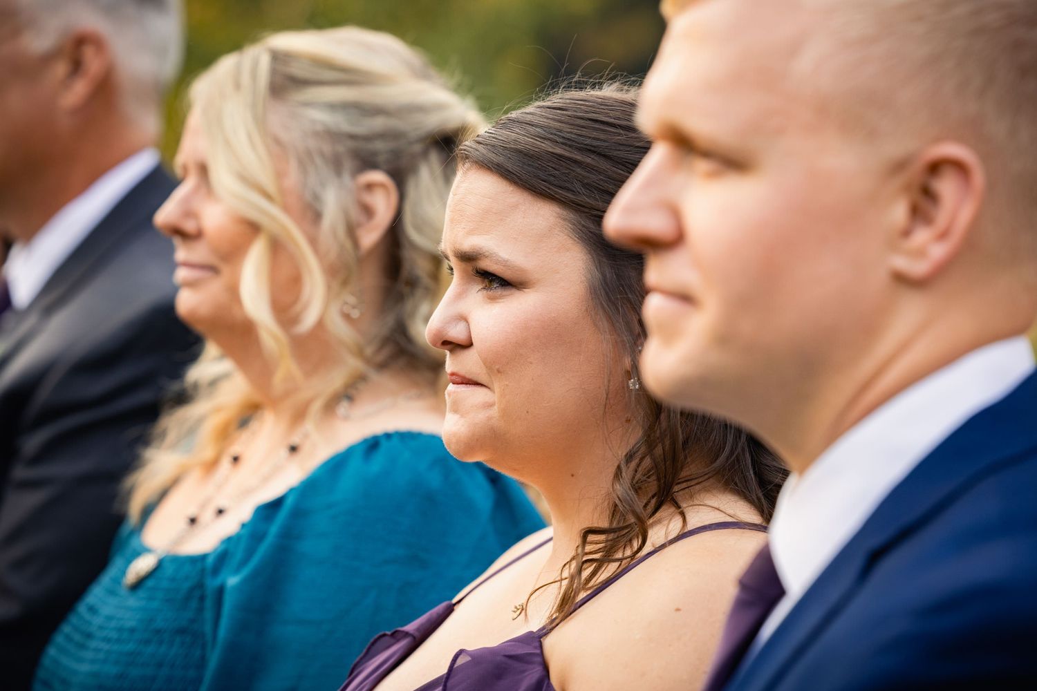 Wedding guests in formal attire smile while seated at an outdoor ceremony.