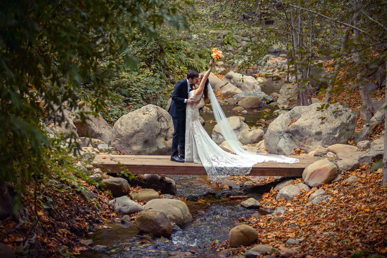 A couple in wedding attire embrace on a wooden footbridge over a rocky stream in a forest setting.