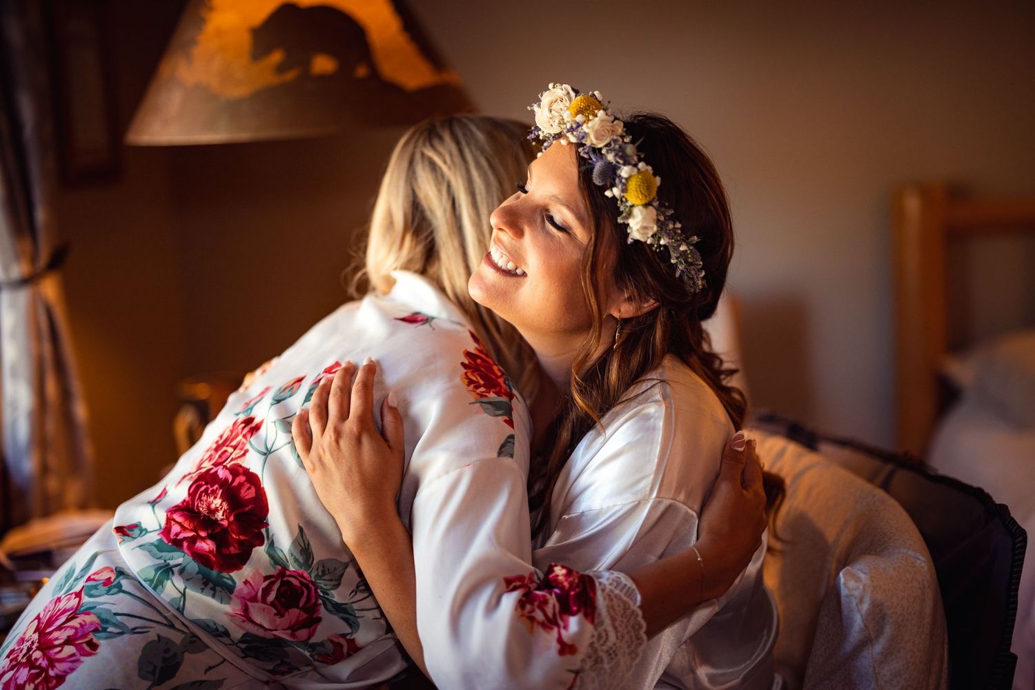 A tender embrace between two people, with one wearing a white embroidered top and floral crown in warm indoor lighting.