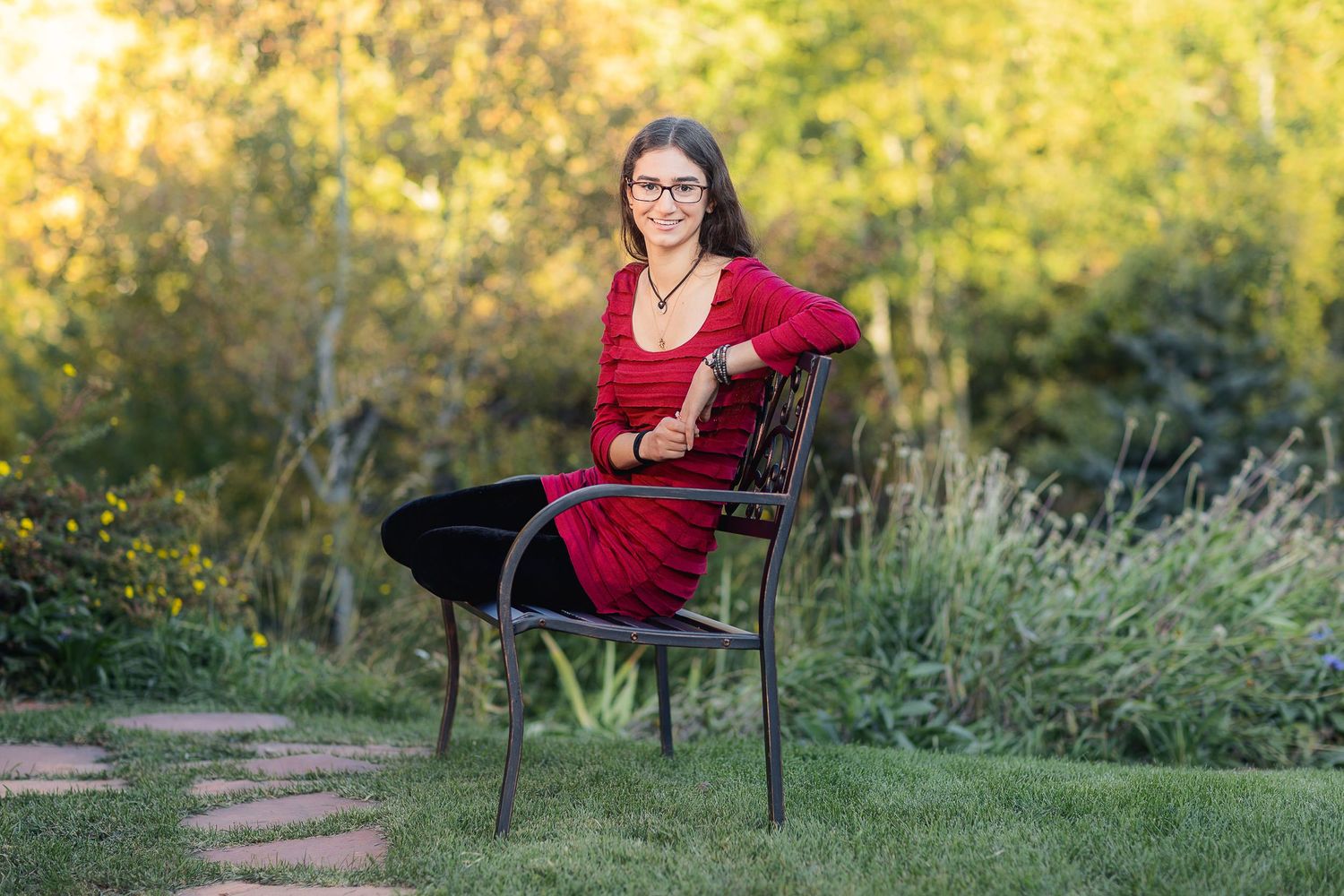 Portrait of someone in red dress seated on black chair in garden setting with golden light.