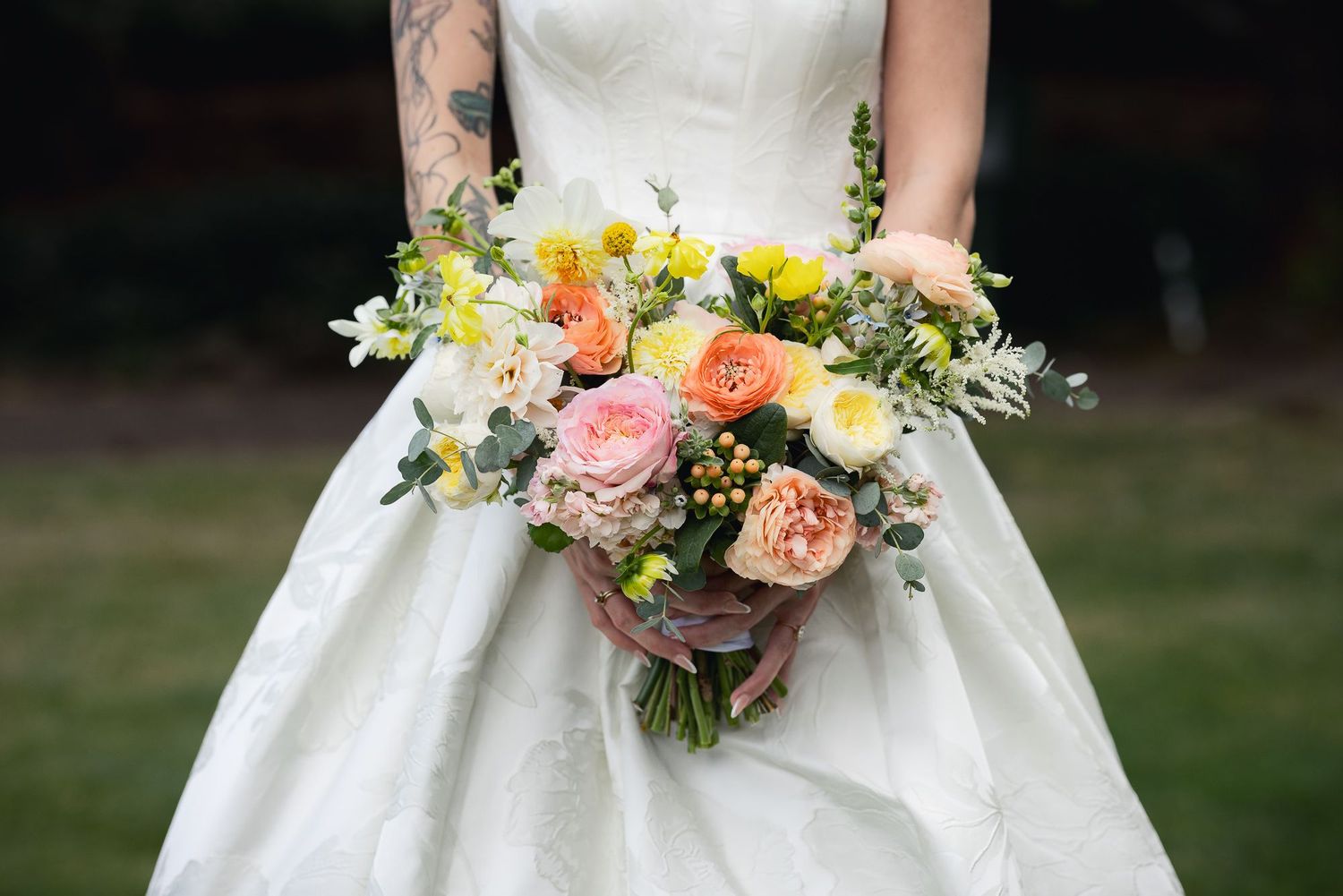 A bridal bouquet featuring pink, peach, yellow and white flowers held against a white wedding dress.