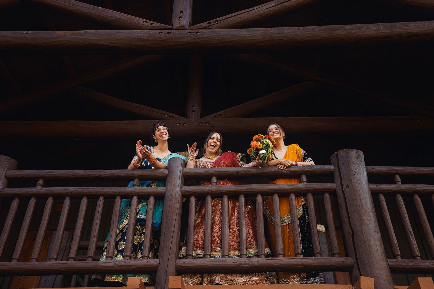 Three women in colorful traditional attire wave and smile from a rustic wooden balcony with log railings.