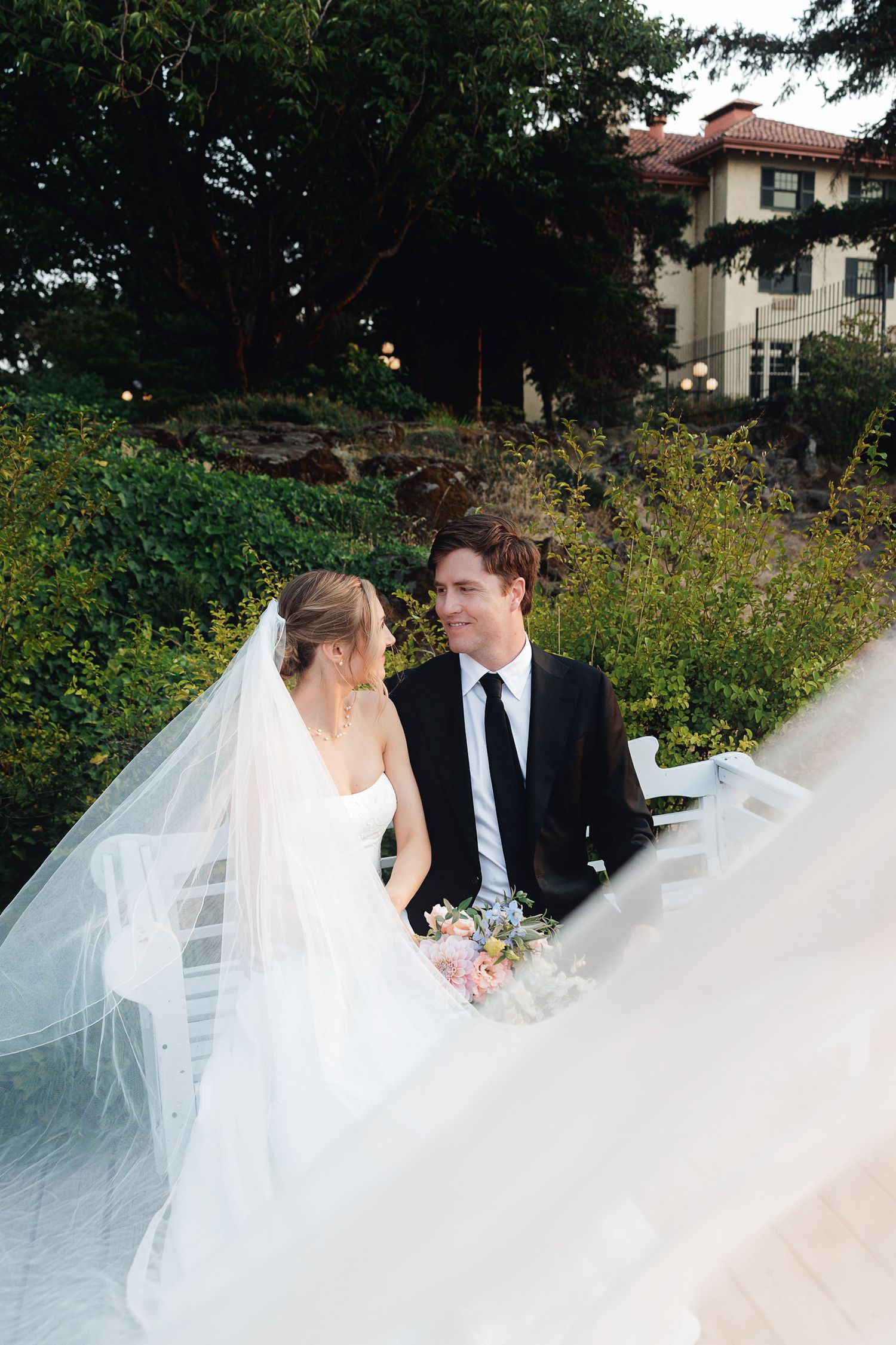 Bride in flowing veil and groom in black suit share a romantic moment outdoors near a vine-covered estate.